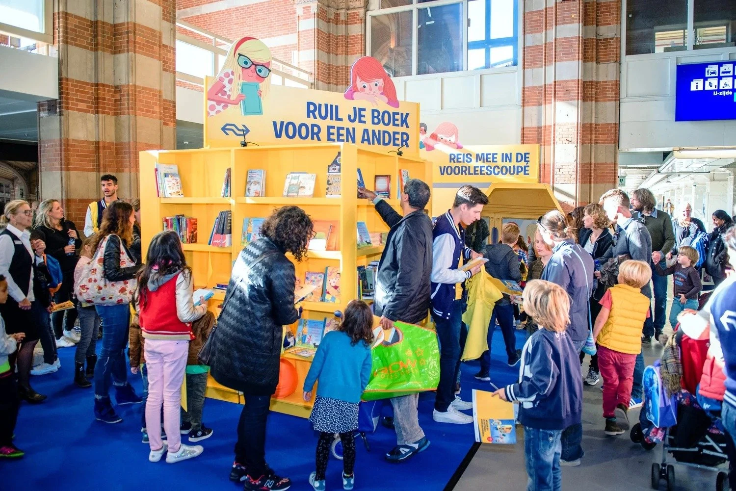 Standontwerp Kinderboekenruil: gele boekenkasten met "Ruil je boek voor een ander", vol kinderen in de stationshal