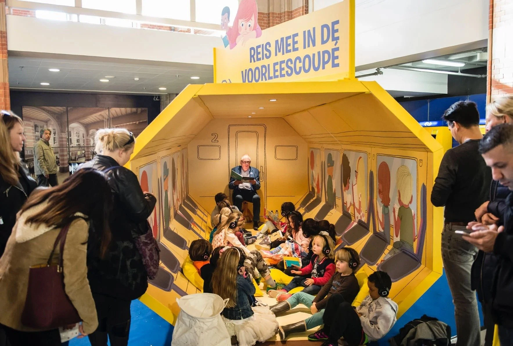 Voorleescoupé in de stand: kinderen zitten in een gele treincoupé en luisteren naar een voorlezer